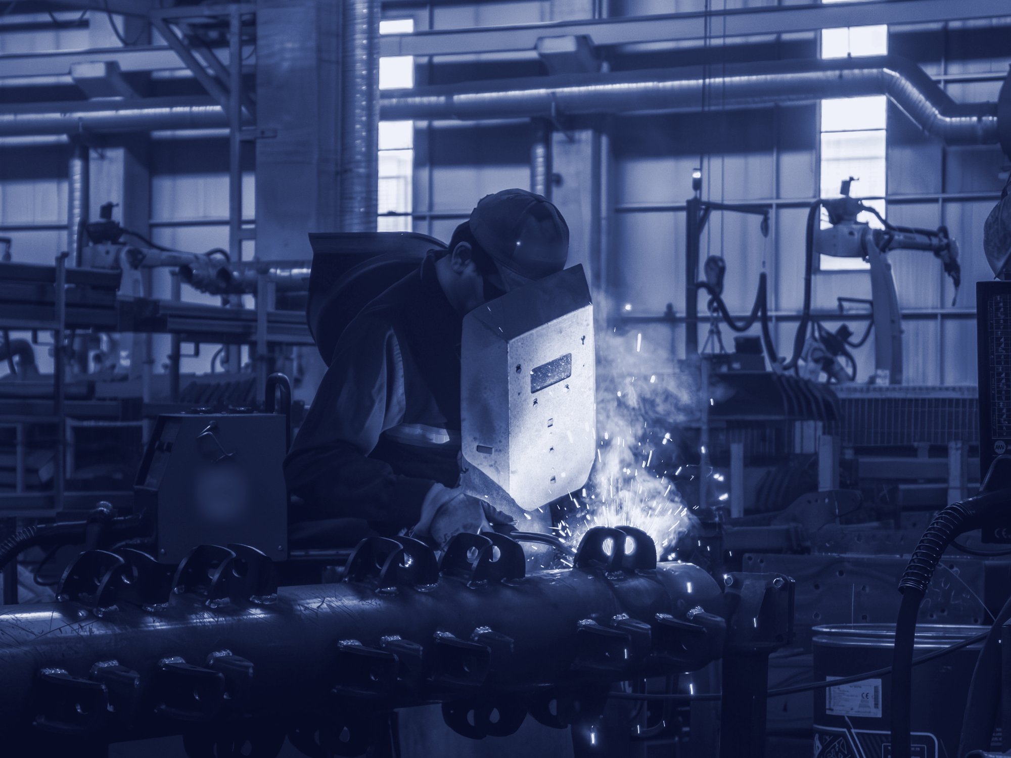 A welder at work on a long fabricated part, with welding robots in the background.